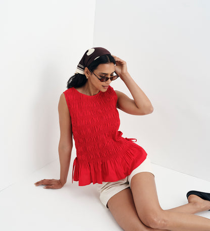 Woman wearing a Malani Red Cotton Poplin Shirred Peplum Shell Top and white shorts on a white background