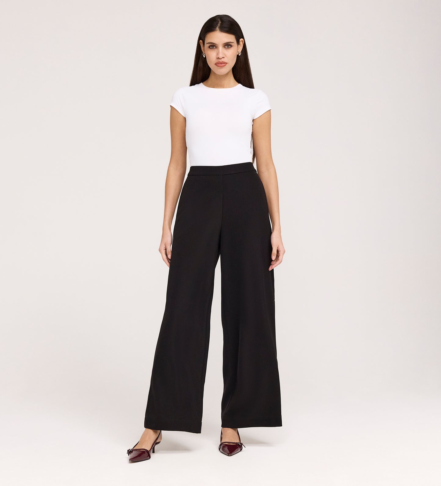 A woman faces forward against a plain background wearing Nat Black Gaberdine Twill Wide Leg Trousers, a fitted white short-sleeve top, and pointed burgundy heels, with her arms relaxed by her sides.