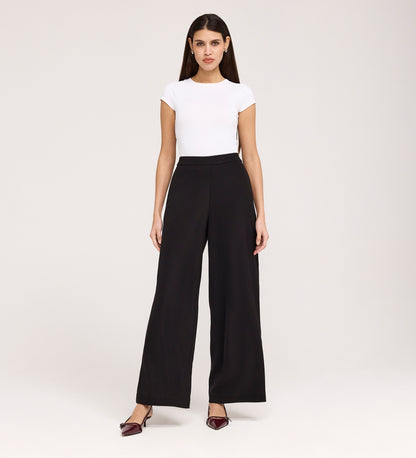 A woman faces forward against a plain background wearing Nat Black Gaberdine Twill Wide Leg Trousers, a fitted white short-sleeve top, and pointed burgundy heels, with her arms relaxed by her sides.