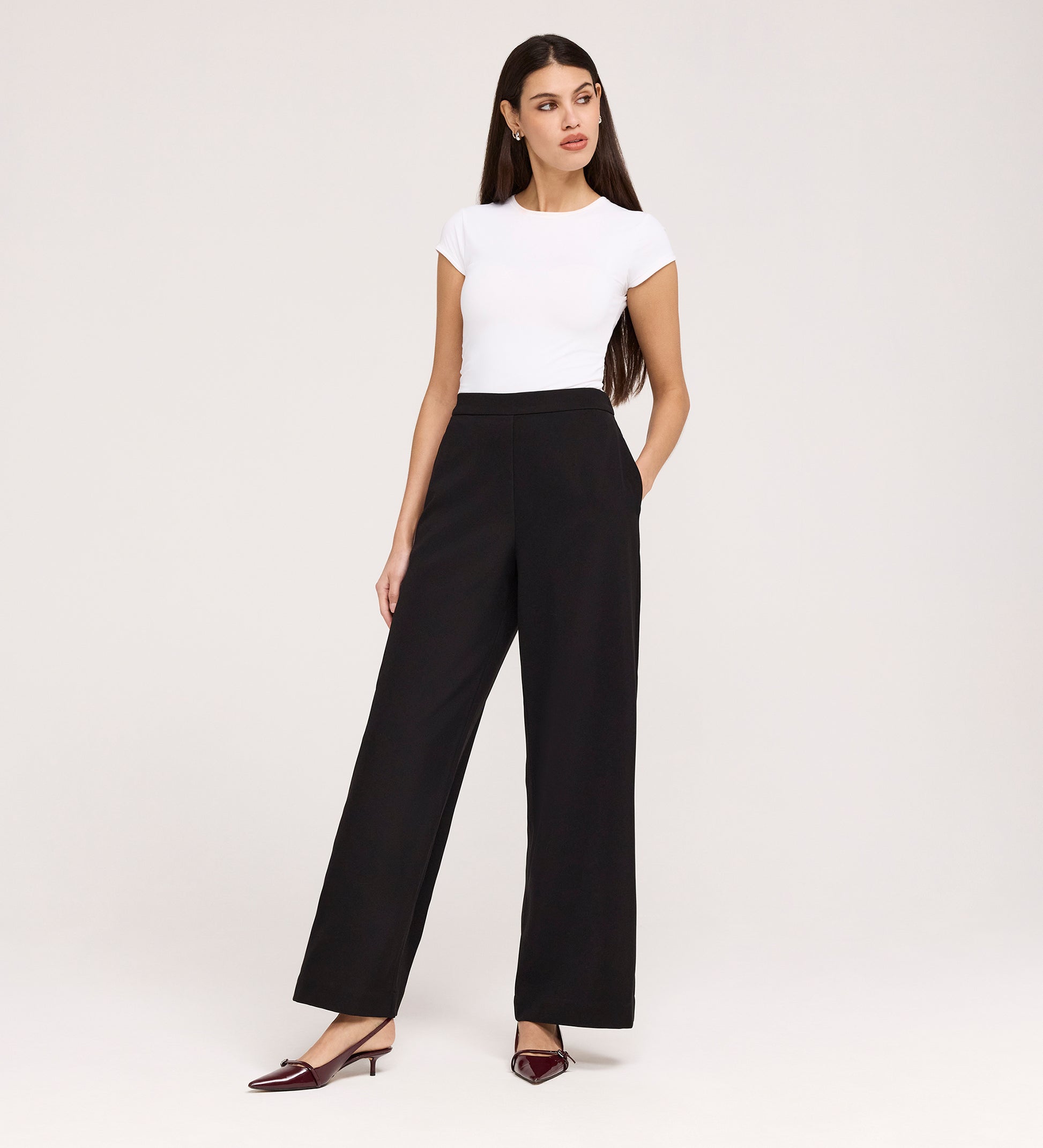 A woman stands against a plain light background wearing the Nat Black Gaberdine Twill Wide Leg Trousers with a fitted white top and burgundy low-heeled shoes, one hand in her pocket as she looks to the side.