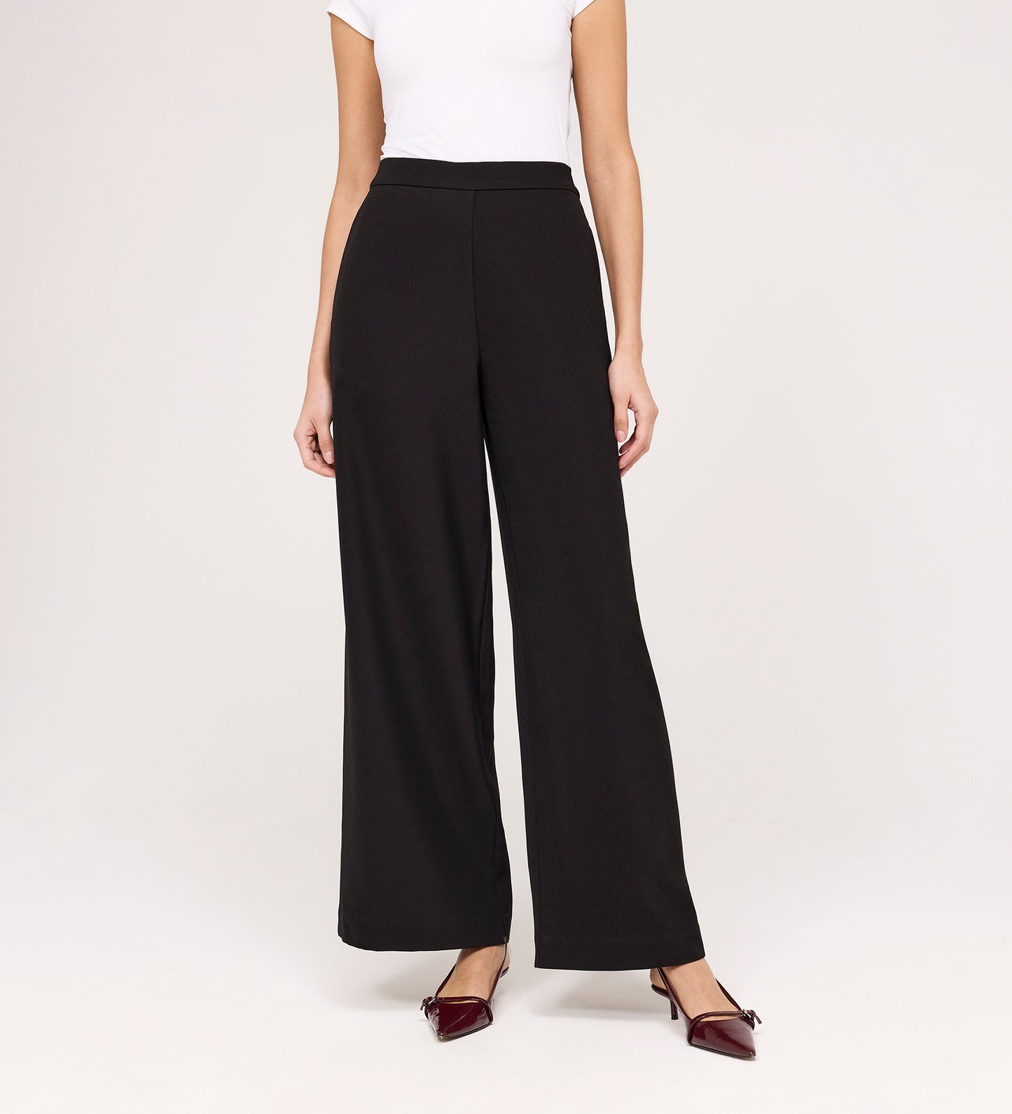 A woman stands against a plain light background wearing a white short-sleeve top, Nat Black Gaberdine Twill Wide Leg Trousers, and burgundy pointed-toe flats. Her face is not visible.