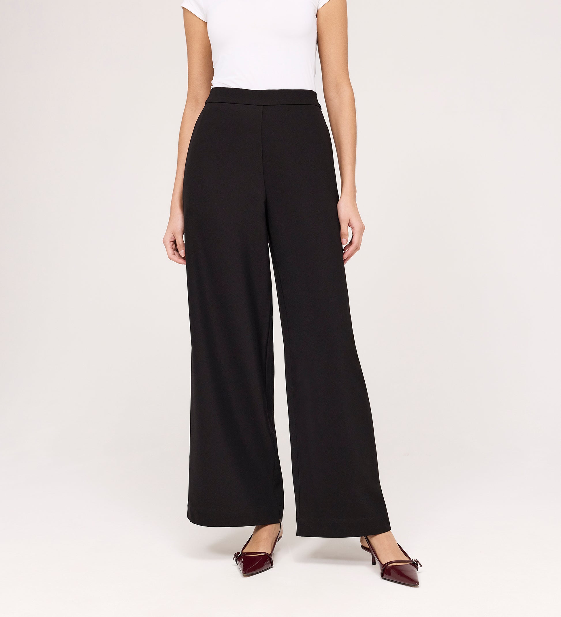 A woman stands against a plain light background wearing a white short-sleeve top, Nat Black Gaberdine Twill Wide Leg Trousers, and burgundy pointed-toe flats. Her face is not visible.