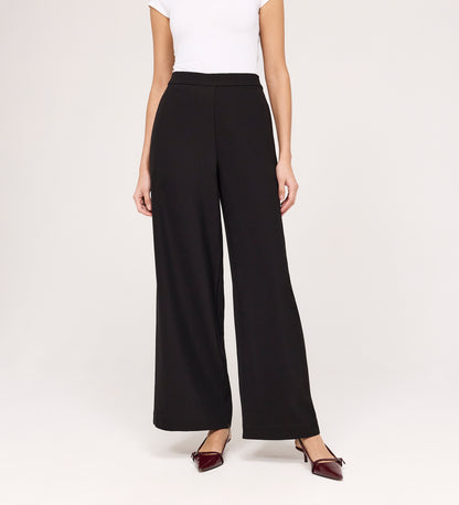 A woman stands against a plain light background wearing a white short-sleeve top, Nat Black Gaberdine Twill Wide Leg Trousers, and burgundy pointed-toe flats. Her face is not visible.