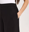 A person models the Nat Black Gaberdine Twill Wide Leg Trousers with a white top, hand in pocket, shown from the waist down against a plain background.