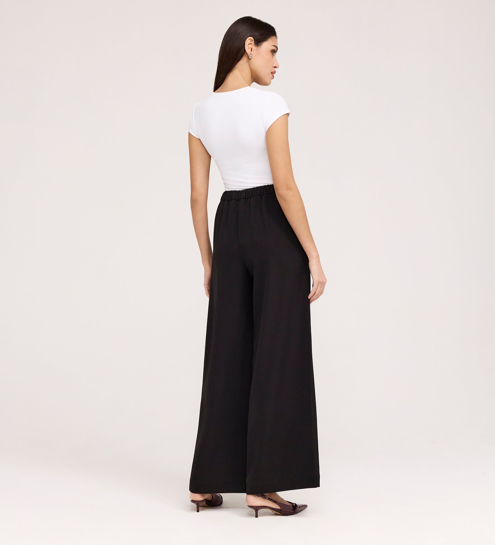 A woman with long dark hair stands against a plain light background, wearing a fitted white short-sleeve top, black flat shoes, and Nat Black Gaberdine Twill Wide Leg Trousers.