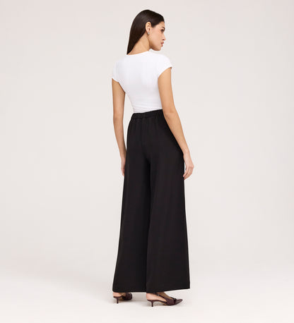 A woman with long dark hair stands against a plain light background, wearing a fitted white short-sleeve top, black flat shoes, and Nat Black Gaberdine Twill Wide Leg Trousers.