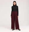 A woman with straight dark hair stands against a plain background, facing forward with a slight smile, wearing a black turtleneck, Nat Burgundy Gaberdine Twill Wide Leg Trousers, and black flats.
