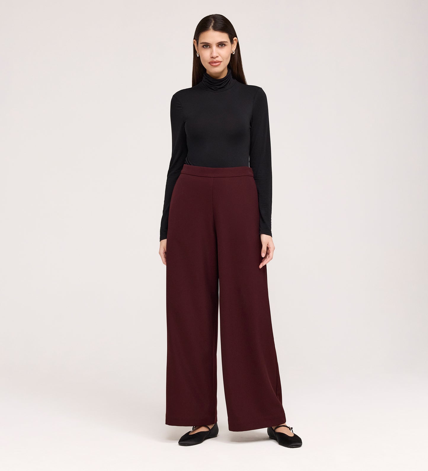 A woman with straight dark hair stands against a plain background, facing forward with a slight smile, wearing a black turtleneck, Nat Burgundy Gaberdine Twill Wide Leg Trousers, and black flats.
