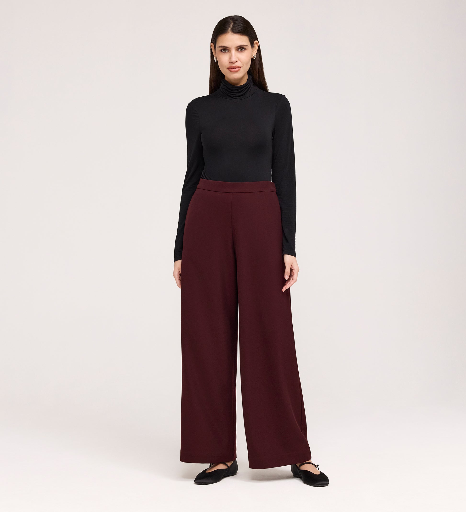 A woman with straight dark hair stands against a plain background, facing forward with a slight smile, wearing a black turtleneck, Nat Burgundy Gaberdine Twill Wide Leg Trousers, and black flats.