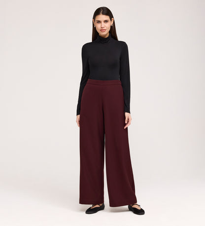 A woman with straight dark hair stands against a plain background, facing forward with a slight smile, wearing a black turtleneck, Nat Burgundy Gaberdine Twill Wide Leg Trousers, and black flats.