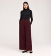 A woman stands with her hands in the pockets of her Nat Burgundy Gaberdine Twill Wide Leg Trousers, paired with a fitted black turtleneck and black flats, against a plain light background.