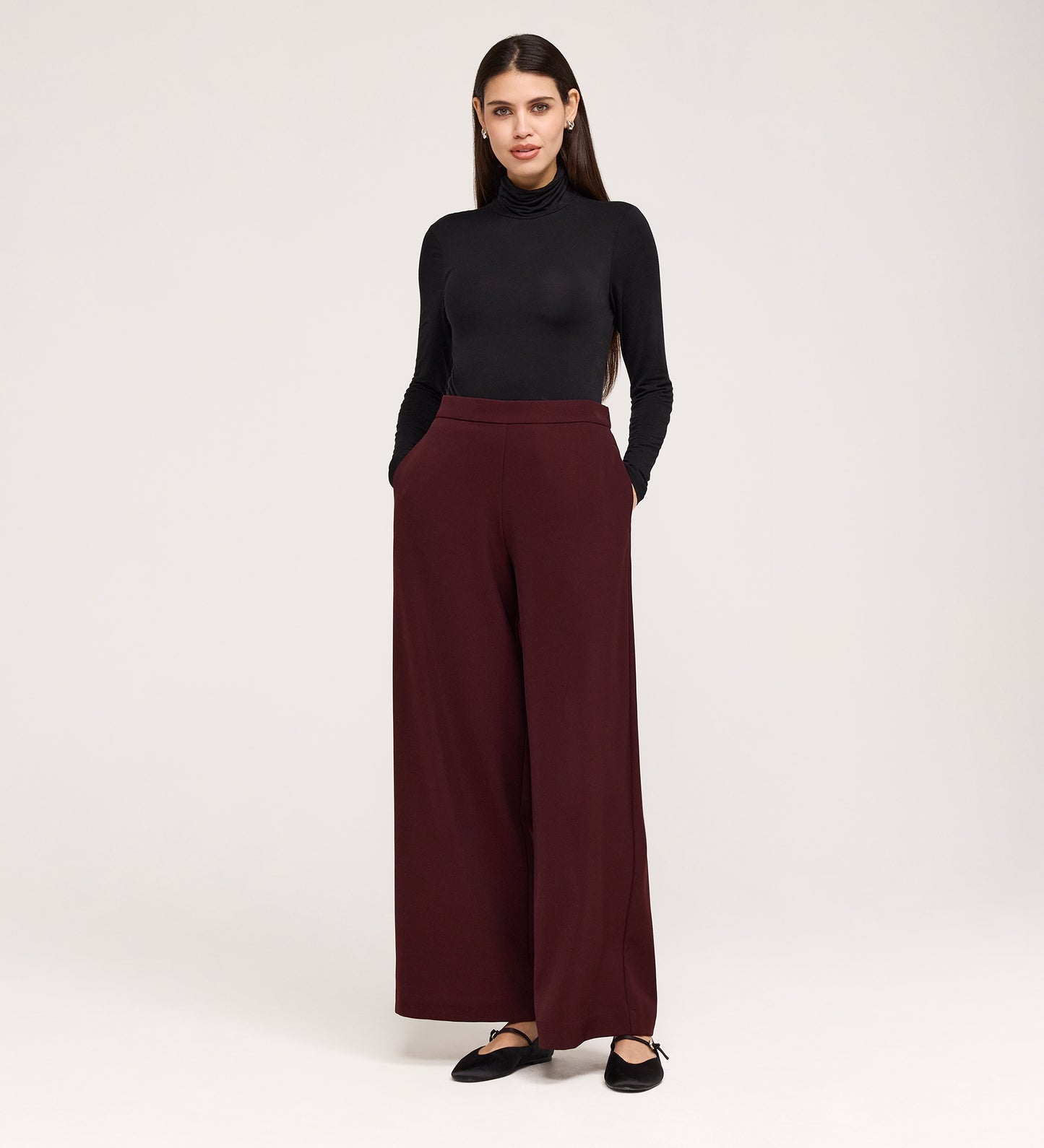 A woman stands with her hands in the pockets of her Nat Burgundy Gaberdine Twill Wide Leg Trousers, paired with a fitted black turtleneck and black flats, against a plain light background.