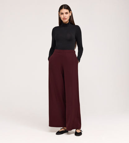 A woman stands with her hands in the pockets of her Nat Burgundy Gaberdine Twill Wide Leg Trousers, paired with a fitted black turtleneck and black flats, against a plain light background.