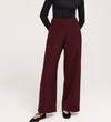 Someone wearing the Nat Burgundy Gaberdine Twill Wide Leg Trousers and a fitted black long-sleeve top stands with hands behind their back against a plain light background.