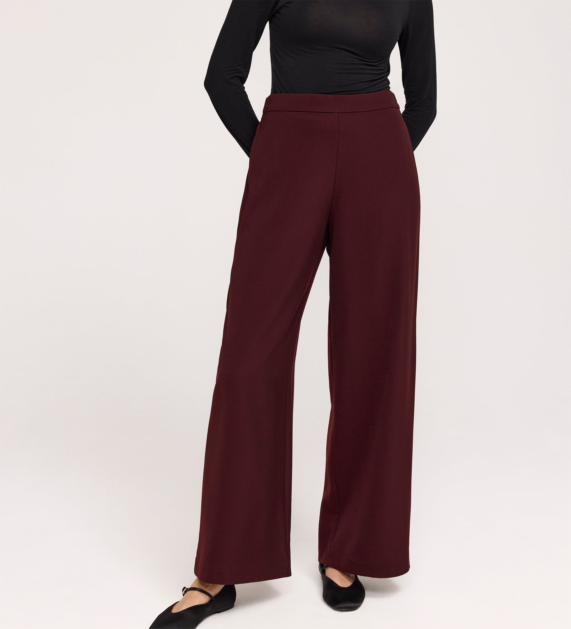 Someone wearing the Nat Burgundy Gaberdine Twill Wide Leg Trousers and a fitted black long-sleeve top stands with hands behind their back against a plain light background.