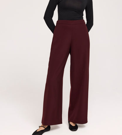Someone wearing the Nat Burgundy Gaberdine Twill Wide Leg Trousers and a fitted black long-sleeve top stands with hands behind their back against a plain light background.