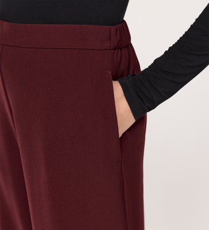 A person in a black long-sleeve top puts their right hand in the pocket of Nat Burgundy Gaberdine Twill Wide Leg Trousers. The background is plain and light-colored.