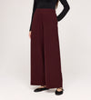 A person models the Nat Burgundy Gaberdine Twill Wide Leg Trousers with a long-sleeved black top and black flat shoes against a plain white background. The person's head is not visible in the image.