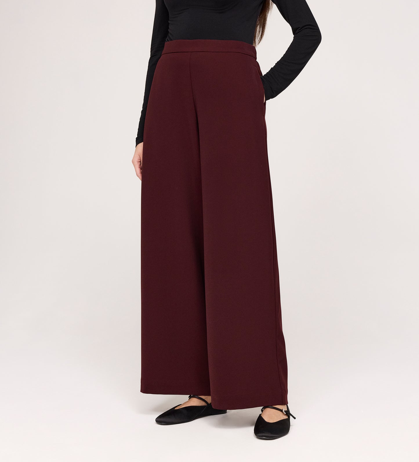 A person models the Nat Burgundy Gaberdine Twill Wide Leg Trousers with a long-sleeved black top and black flat shoes against a plain white background. The person's head is not visible in the image.