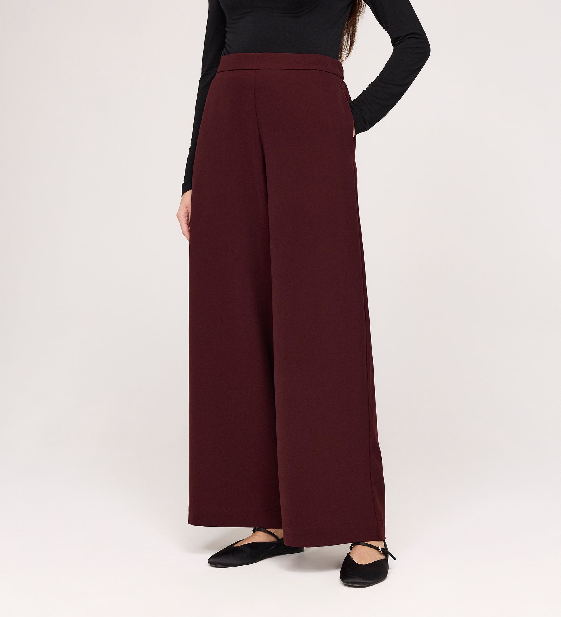 A person models the Nat Burgundy Gaberdine Twill Wide Leg Trousers with a long-sleeved black top and black flat shoes against a plain white background. The person's head is not visible in the image.