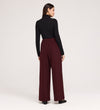 A woman stands with her back to the camera, wearing black long sleeves and the Nat Burgundy Gaberdine Twill Wide Leg Trousers with black shoes, set against a plain light background.