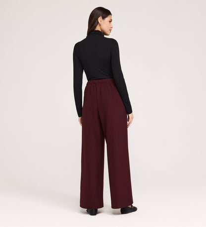 A woman stands with her back to the camera, wearing black long sleeves and the Nat Burgundy Gaberdine Twill Wide Leg Trousers with black shoes, set against a plain light background.
