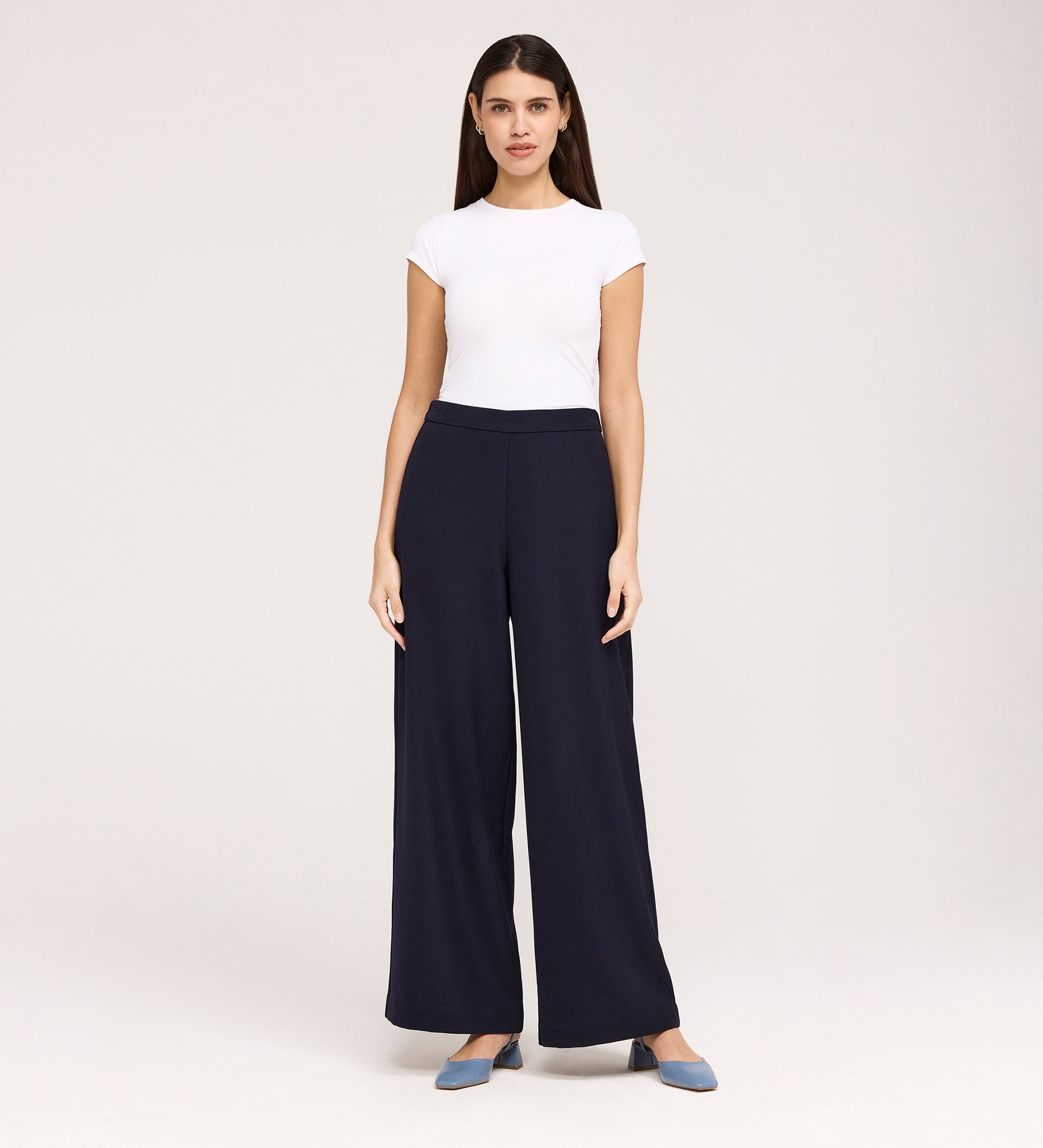 A woman stands facing forward, wearing a white short-sleeve top, Nat Navy Gaberdine Twill Wide Leg Trousers, and blue flats against a plain light background.