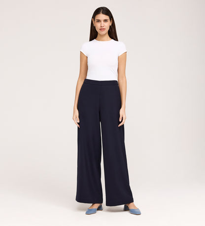 A woman stands facing forward, wearing a white short-sleeve top, Nat Navy Gaberdine Twill Wide Leg Trousers, and blue flats against a plain light background.