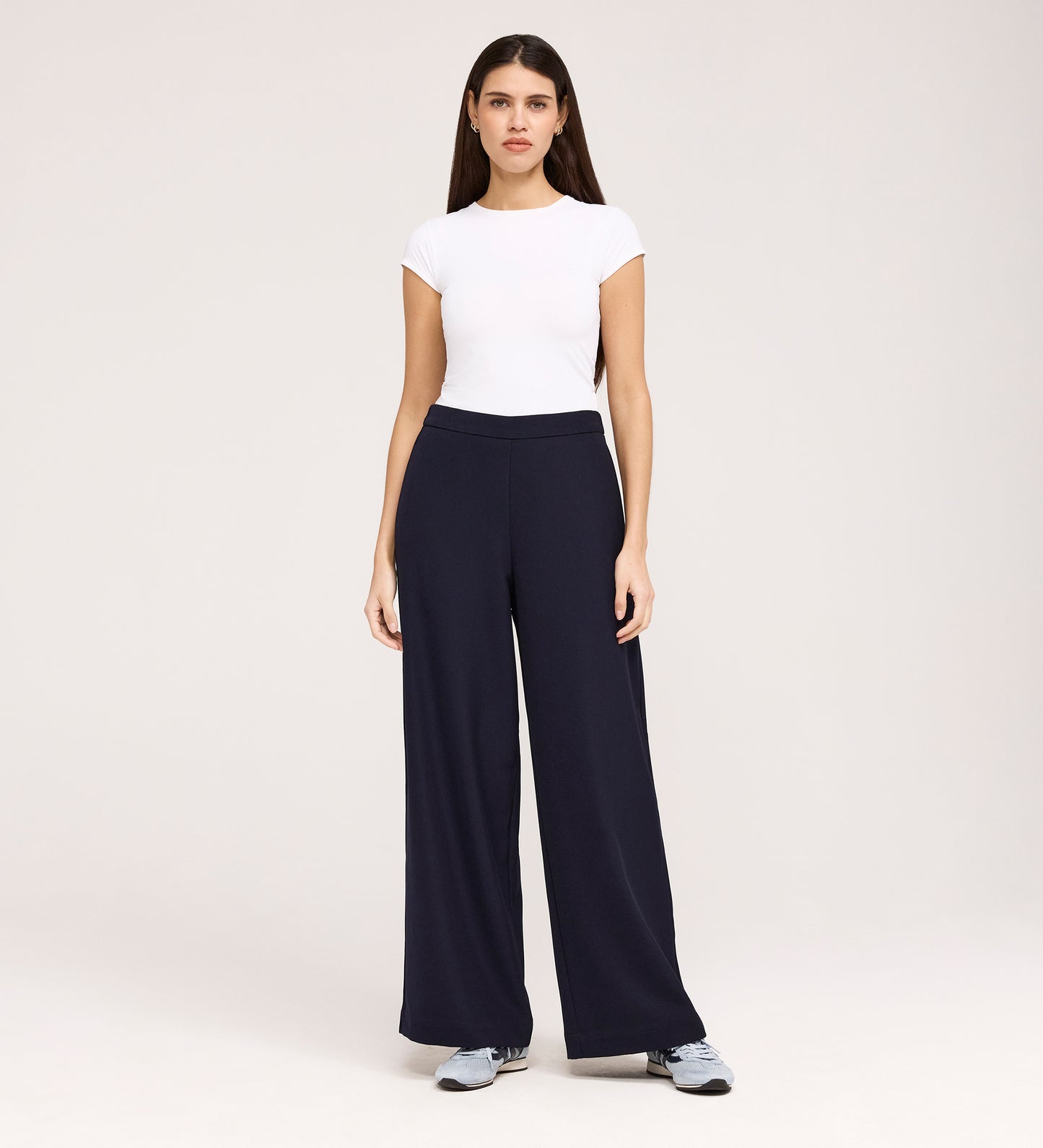 A woman faces forward against a plain light background, wearing a fitted white short-sleeve top, gray sneakers, and the Nat Navy Gaberdine Twill Wide Leg Trousers.