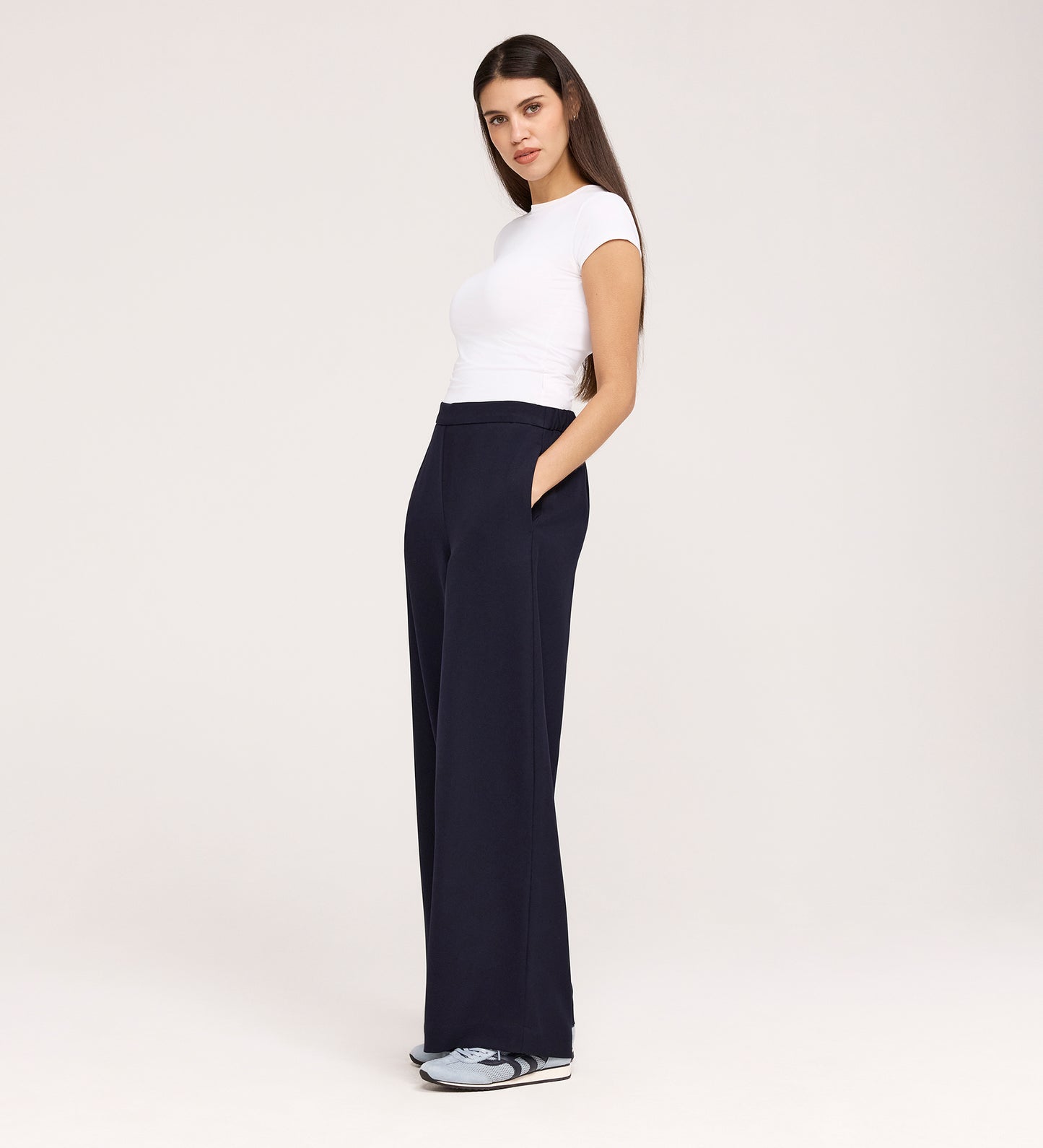 A woman stands with her hands in the pockets of her Nat Navy Gaberdine Twill Wide Leg Trousers, wearing a fitted white t-shirt and sneakers. She is facing slightly to the side, framed by long dark hair and a neutral background.