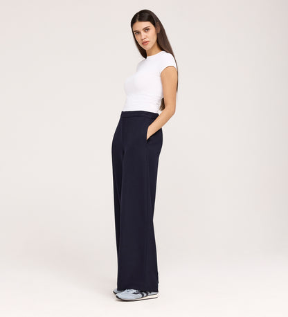 A woman stands with her hands in the pockets of her Nat Navy Gaberdine Twill Wide Leg Trousers, wearing a fitted white t-shirt and sneakers. She is facing slightly to the side, framed by long dark hair and a neutral background.