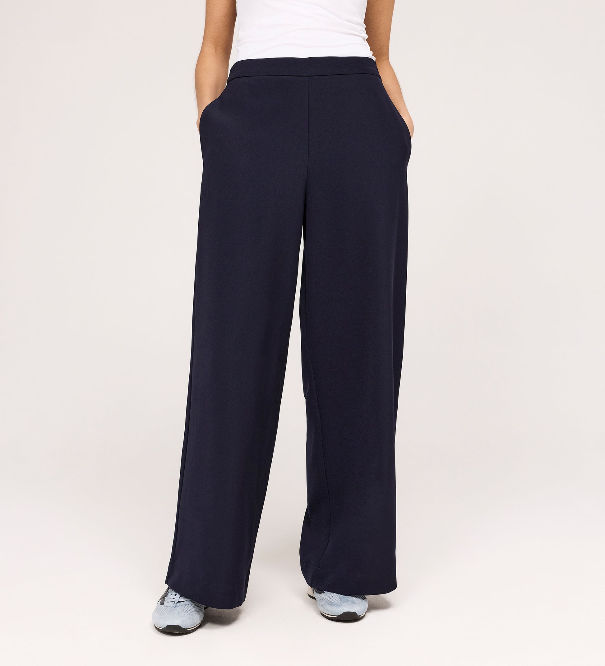 Wearing the Nat Navy Gaberdine Twill Wide Leg Trousers, a person with hands in pockets, white top, and gray sneakers stands against a plain light background.