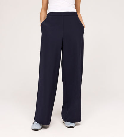 Wearing the Nat Navy Gaberdine Twill Wide Leg Trousers, a person with hands in pockets, white top, and gray sneakers stands against a plain light background.