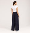 Woman stands with her back to the camera, wearing a fitted white short-sleeve top, sneakers, and Nat Navy Gaberdine Twill Wide Leg Trousers against a plain light background.