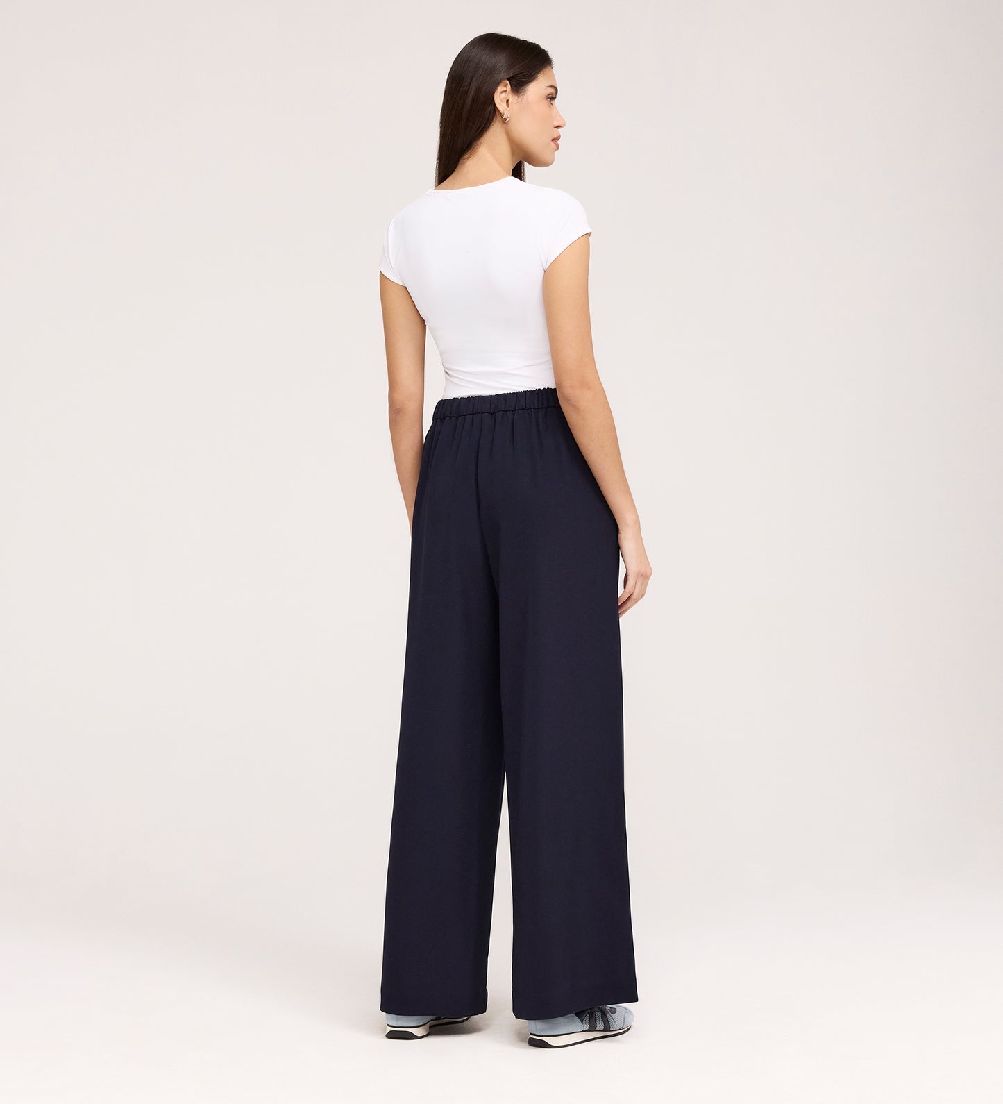 Woman stands with her back to the camera, wearing a fitted white short-sleeve top, sneakers, and Nat Navy Gaberdine Twill Wide Leg Trousers against a plain light background.