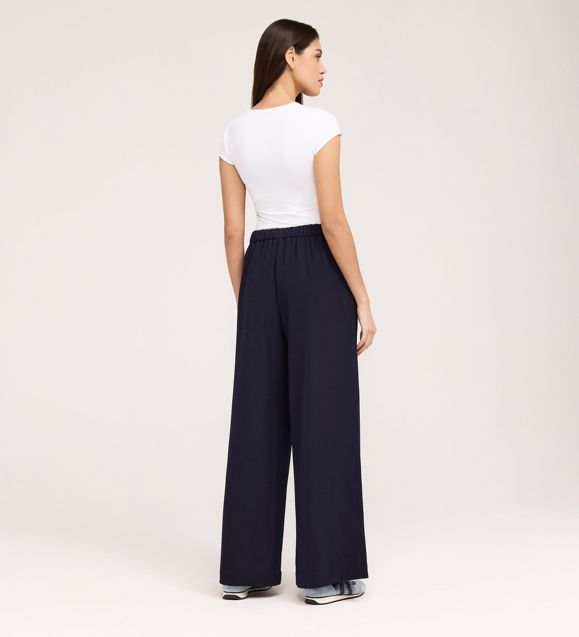 Woman stands with her back to the camera, wearing a fitted white short-sleeve top, sneakers, and Nat Navy Gaberdine Twill Wide Leg Trousers against a plain light background.