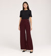 A woman stands against a plain background, wearing a black short-sleeve top and the Nellie Burgundy Gaberdine Twill Wide Leg Trousers with decorative side buttons—an elegant staple—paired with black heels.