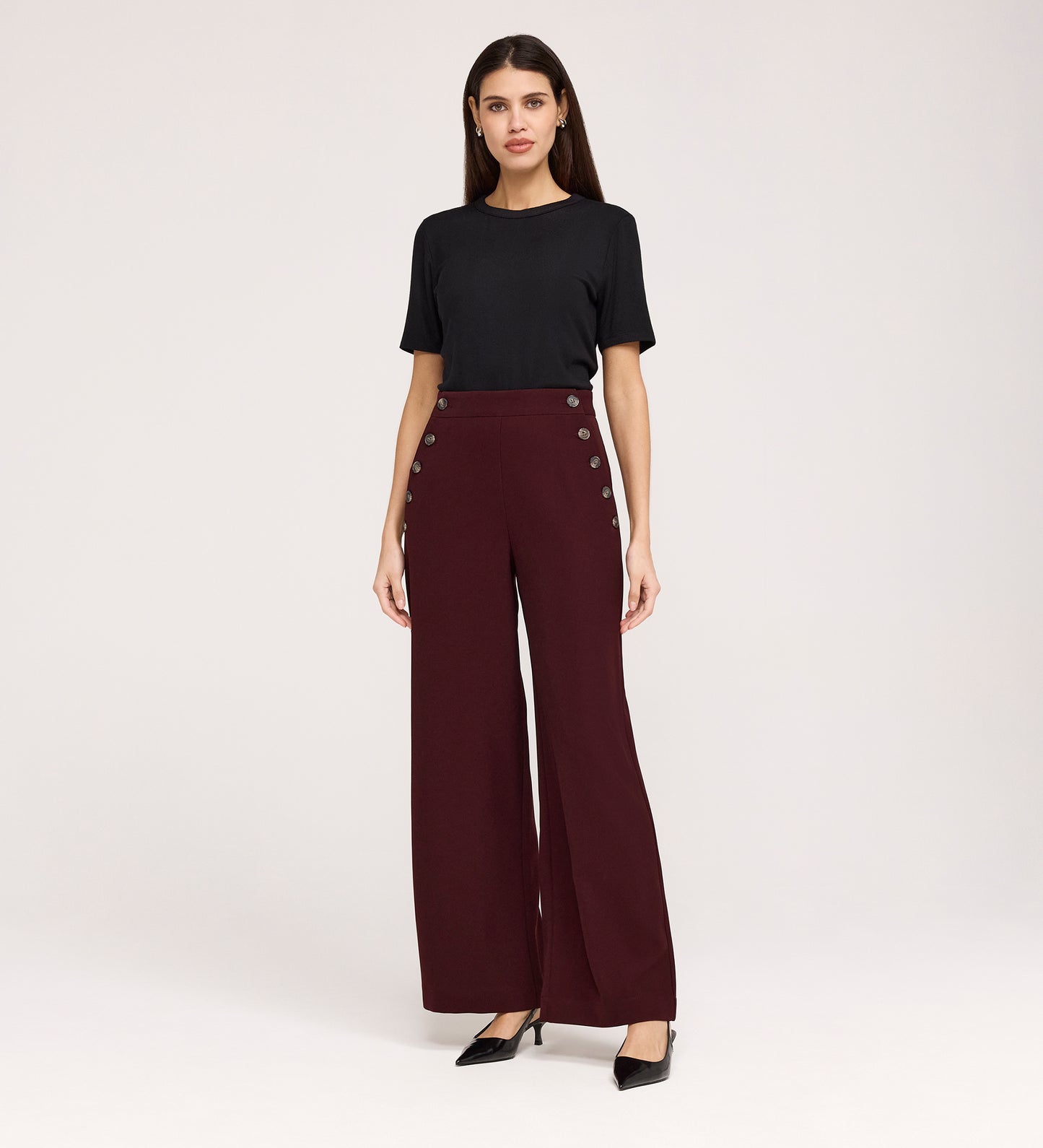 A woman stands against a plain background, wearing a black short-sleeve top and the Nellie Burgundy Gaberdine Twill Wide Leg Trousers with decorative side buttons—an elegant staple—paired with black heels.