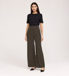 A woman poses against a plain background wearing a black short-sleeve top, black heels, and Nellie Mushroom Gaberdine Twill Wide Leg Trousers with decorative side buttons.