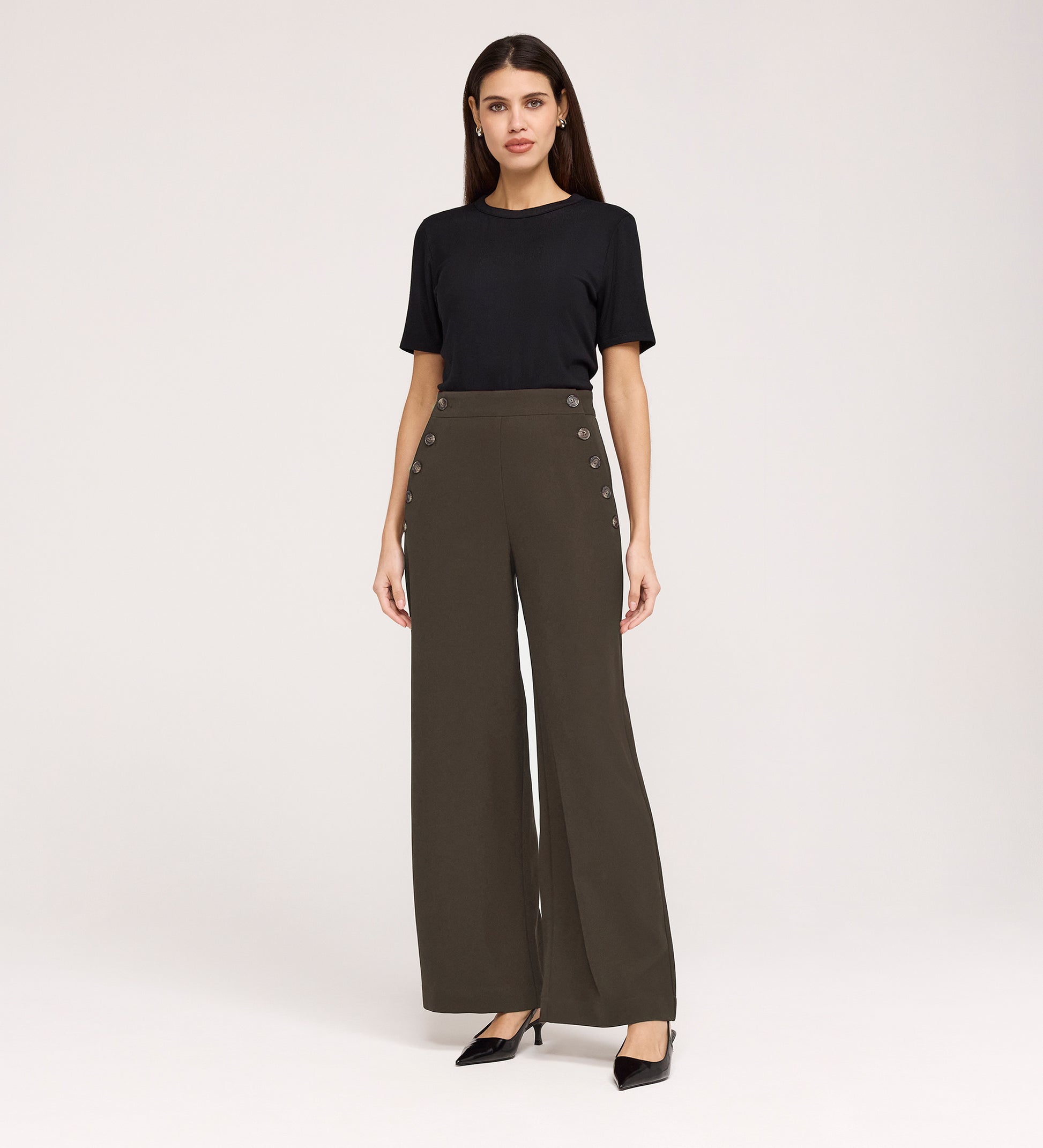 A woman poses against a plain background wearing a black short-sleeve top, black heels, and Nellie Mushroom Gaberdine Twill Wide Leg Trousers with decorative side buttons.