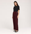 A woman with long dark hair and a neutral expression stands against a light background, wearing the Nellie Burgundy Gaberdine Twill Wide Leg Trousers, a black short-sleeve top, and black pointed-toe heels.
