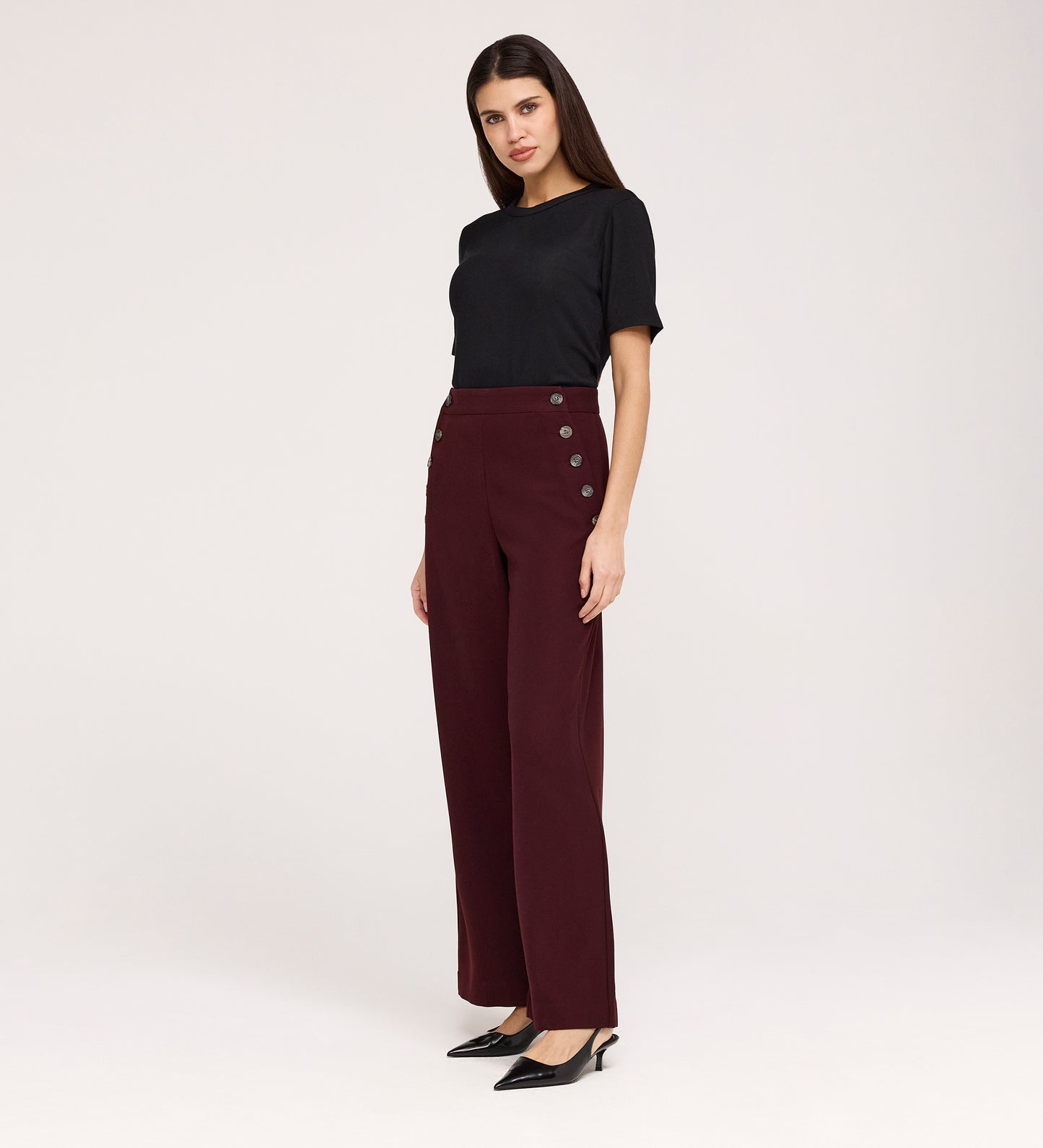 A woman with long dark hair and a neutral expression stands against a light background, wearing the Nellie Burgundy Gaberdine Twill Wide Leg Trousers, a black short-sleeve top, and black pointed-toe heels.