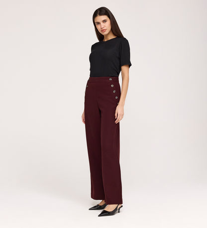 A woman with long dark hair and a neutral expression stands against a light background, wearing the Nellie Burgundy Gaberdine Twill Wide Leg Trousers, a black short-sleeve top, and black pointed-toe heels.