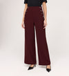 A person stands against a light background wearing Nellie Burgundy Gaberdine Twill Wide Leg Trousers with double rows of decorative buttons, paired with a black top and black pointed-toe heels.