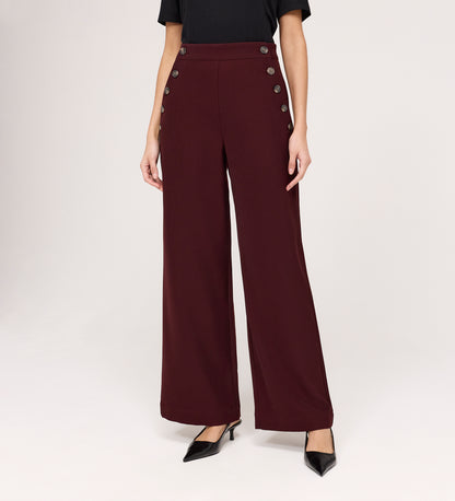 A person stands against a light background wearing Nellie Burgundy Gaberdine Twill Wide Leg Trousers with double rows of decorative buttons, paired with a black top and black pointed-toe heels.