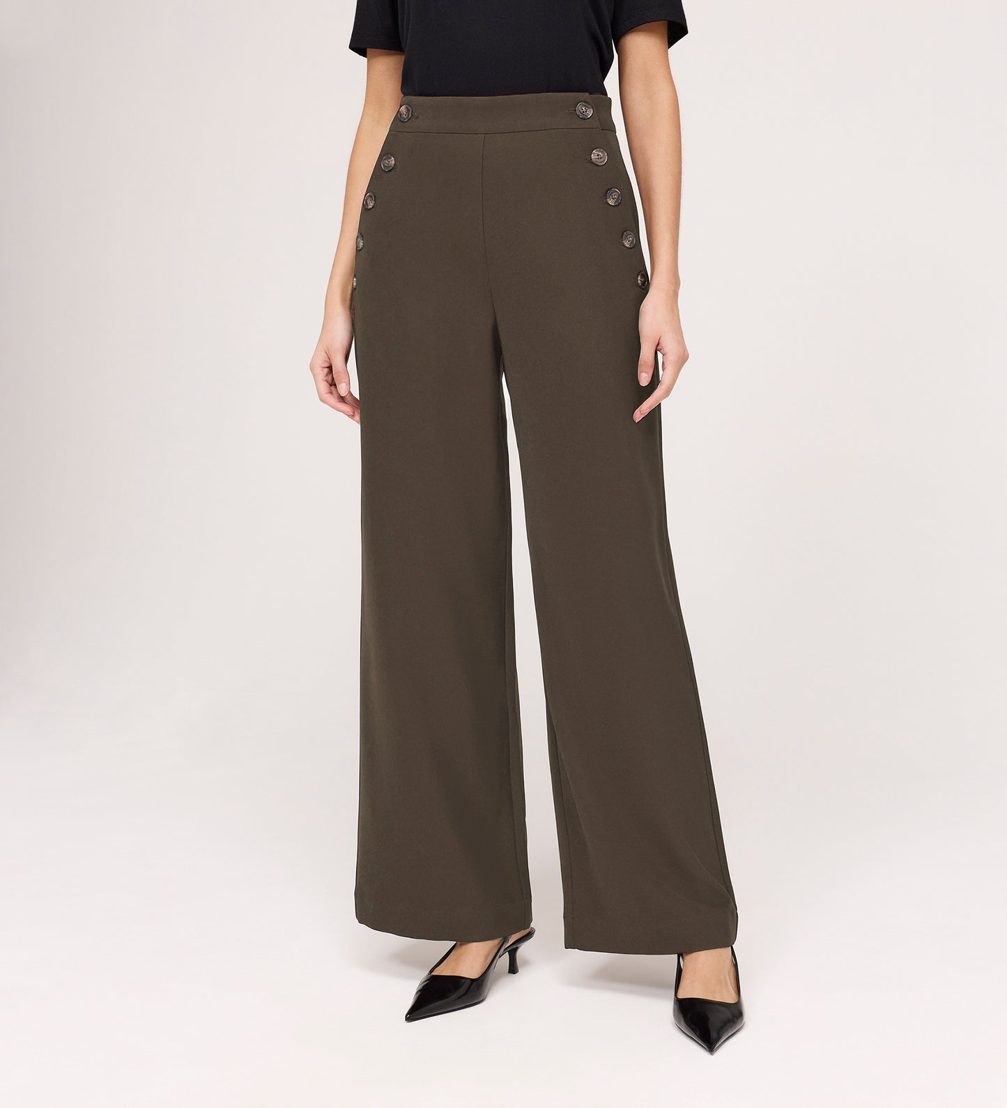 Wearing the Nellie Mushroom Gaberdine Twill Wide Leg Trousers with decorative side buttons, a black short-sleeve top, and black pointed-toe heels, a person stands against a plain background.