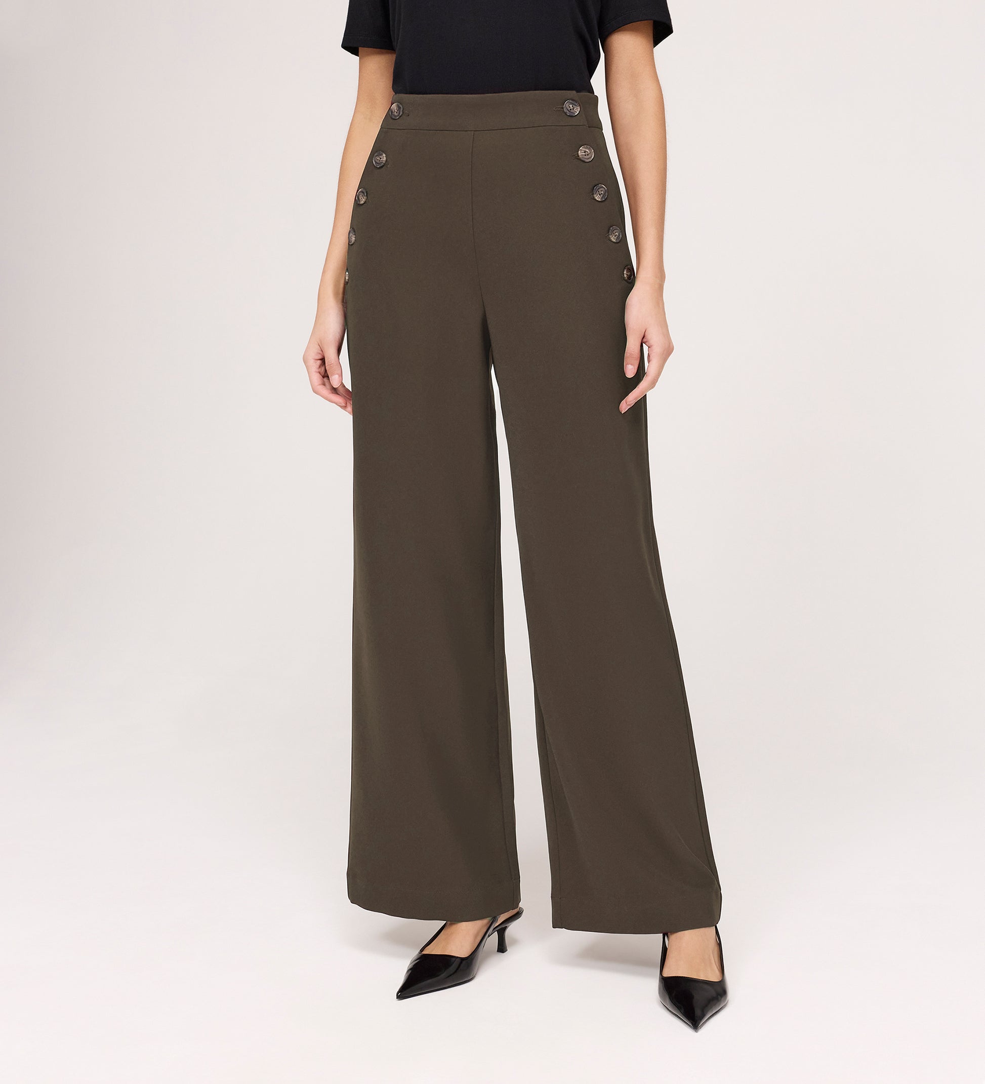 Wearing the Nellie Mushroom Gaberdine Twill Wide Leg Trousers with decorative side buttons, a black short-sleeve top, and black pointed-toe heels, a person stands against a plain background.