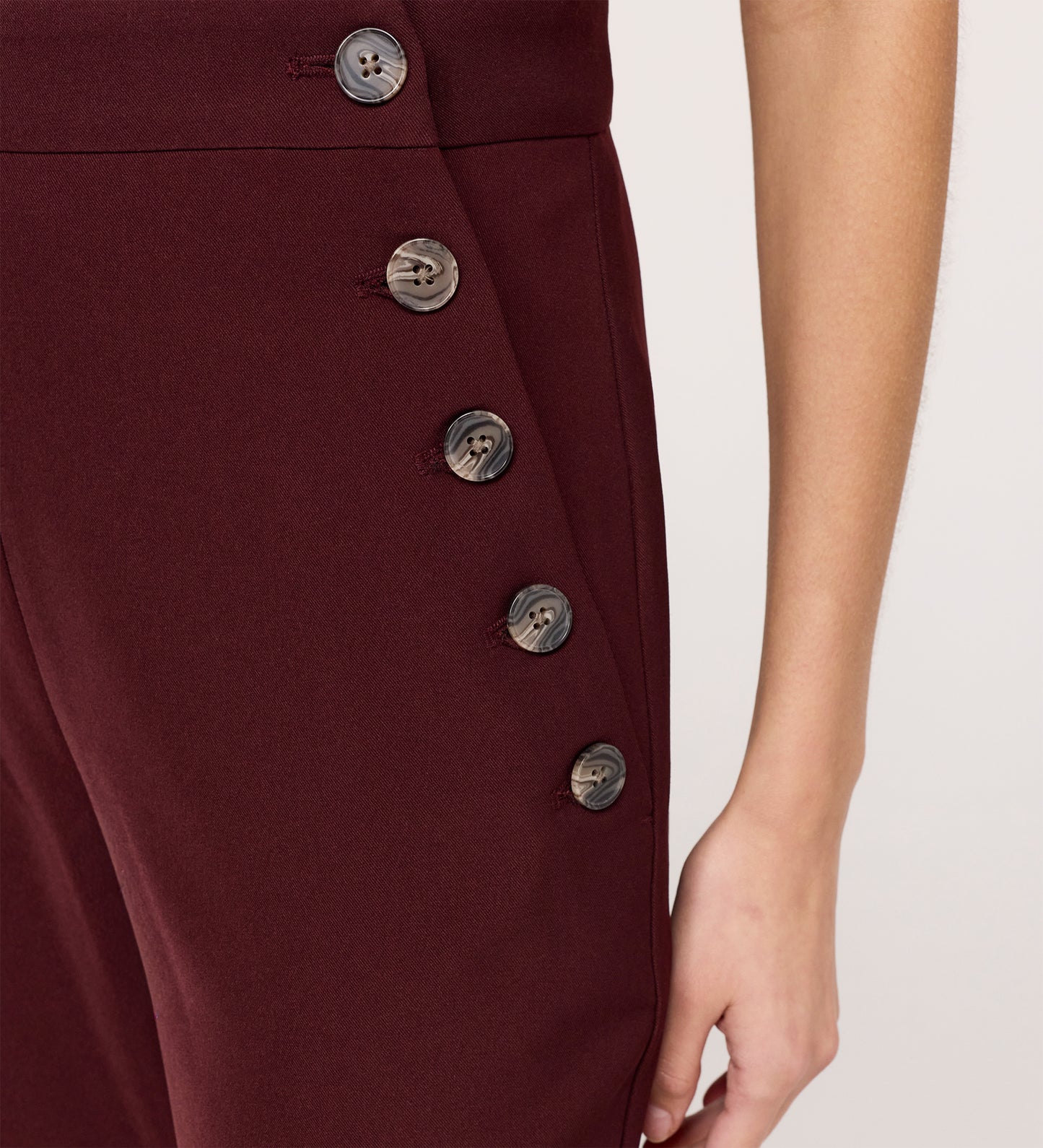 Close-up of a person wearing the Nellie Burgundy Gaberdine Twill Wide Leg Trousers featuring five dark side buttons; only the lower torso and right arm are shown.