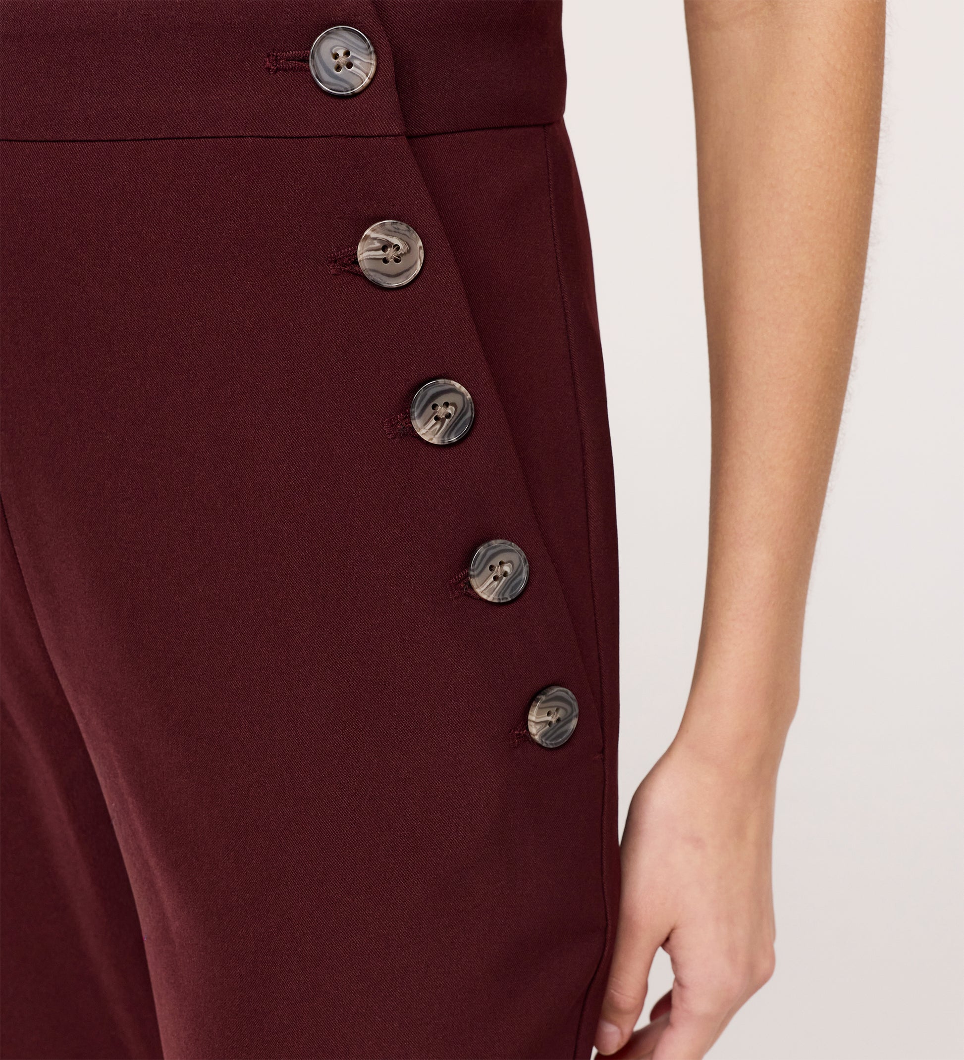 Close-up of a person wearing the Nellie Burgundy Gaberdine Twill Wide Leg Trousers featuring five dark side buttons; only the lower torso and right arm are shown.