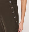 A close-up of a person wearing Nellie Mushroom Gaberdine Twill Wide Leg Trousers, high-waisted with a dark olive green hue and side row of black buttons. Their right arm and hand rest at their side.
